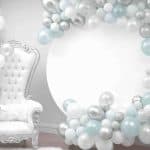 A white ornate chair with a teddy bear sits beside blue and white balloons. A large silver balloon arch decorates the background, perfect for capturing memories at a NY party hall in Queens. The scene is soft and elegant for any celebration.