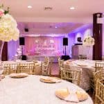 Elegant banquet hall decorated for a formal event, with round tables covered in white floral linens, gold chairs, tall floral centerpieces, and a draped stage at the front, all under soft pink and purple lighting.